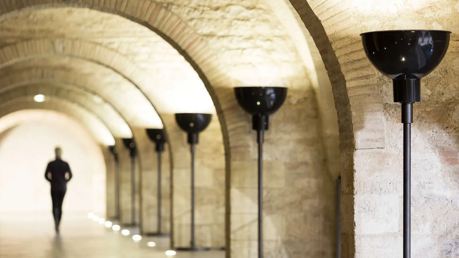 Underground arched corridor with stone walls illuminated by black upright lamps, a silhouetted figure visible in the distance