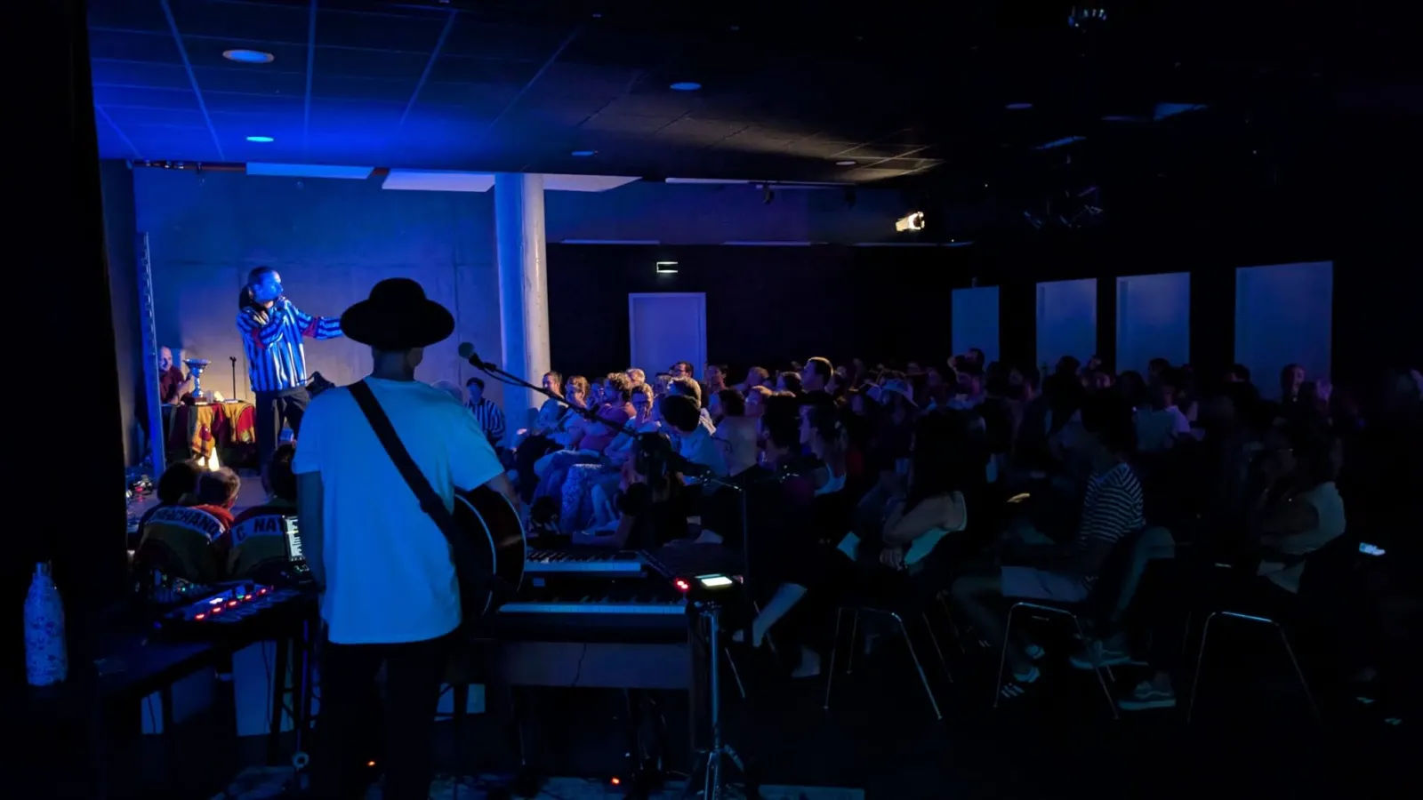 Live music performance at L'Estrade in Bordeaux with a musician wearing a wide-brimmed hat playing guitar on stage, addressing an seated audience in a blue-lit indoor venue with large windows.