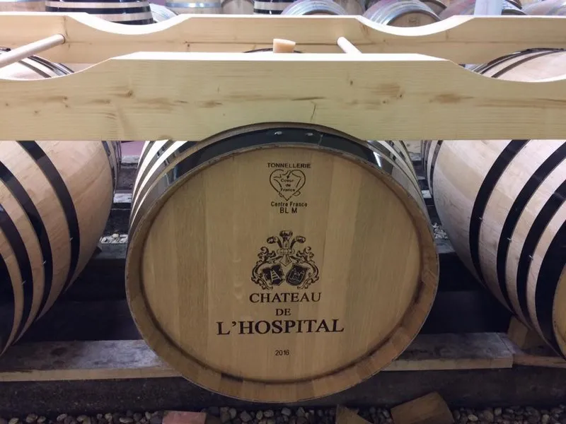 Wooden wine barrel labeled 'Château de l'Hospital' with vintage 2018, displayed in a temperature-controlled cellar among other barrels with metal bands