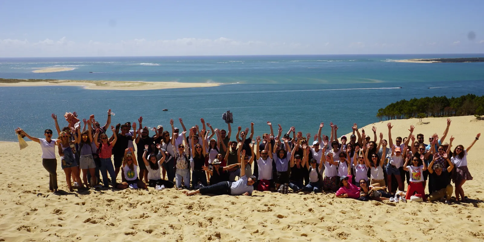 Large group of people with raised arms celebrating on a sandy beach with turquoise sea and sand dunes in the background