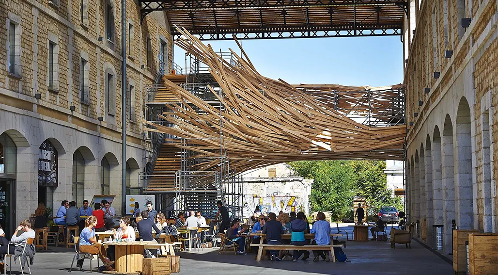 Outdoor courtyard in a historic stone building complex with visitors seated at wooden tables under a woven wooden canopy structure, featuring arches and modern architectural elements.