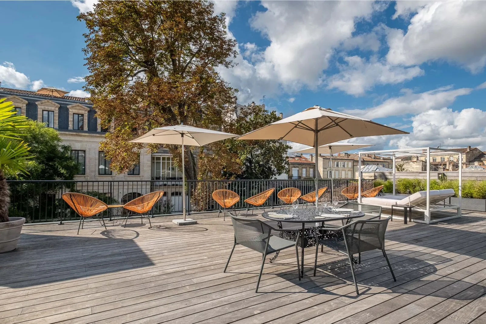 Rooftop terrace of Palais Gallien Hotel & Spa in Bordeaux with orange modern chairs, dining tables with umbrellas, wooden deck, and panoramic views of the city's historic architecture under a blue sky.