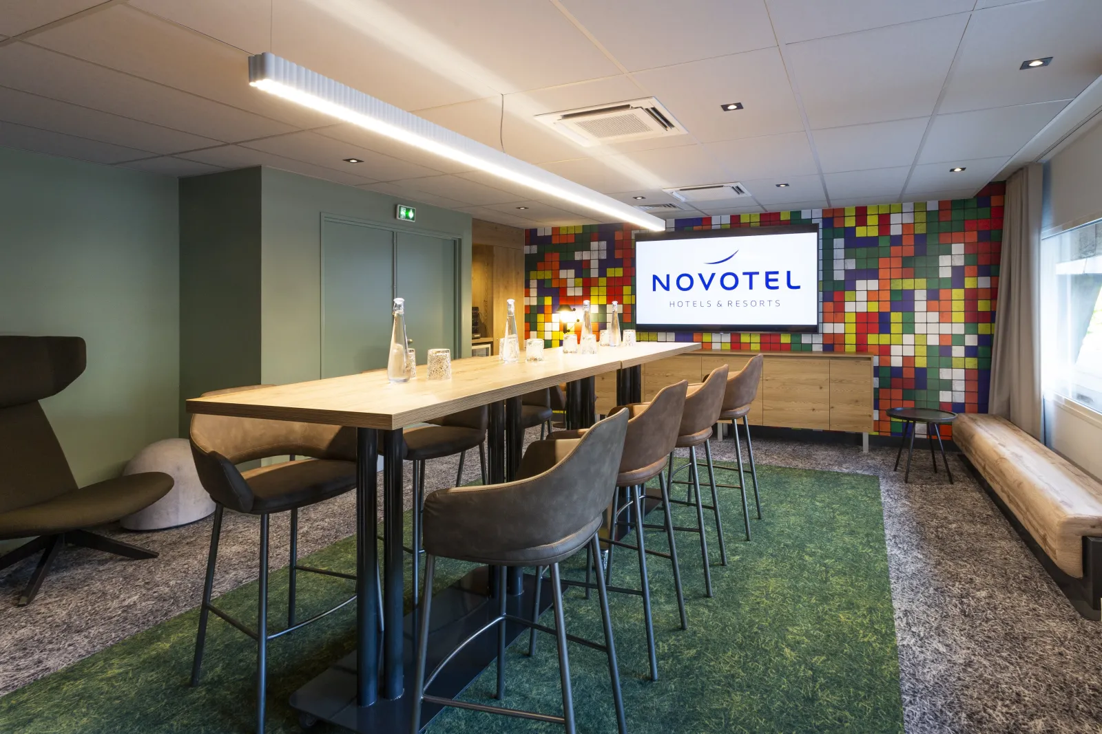 Modern meeting room at Novotel Bordeaux Centre with high-top table, bar seating, colorful mosaic wall feature, and contemporary lighting.