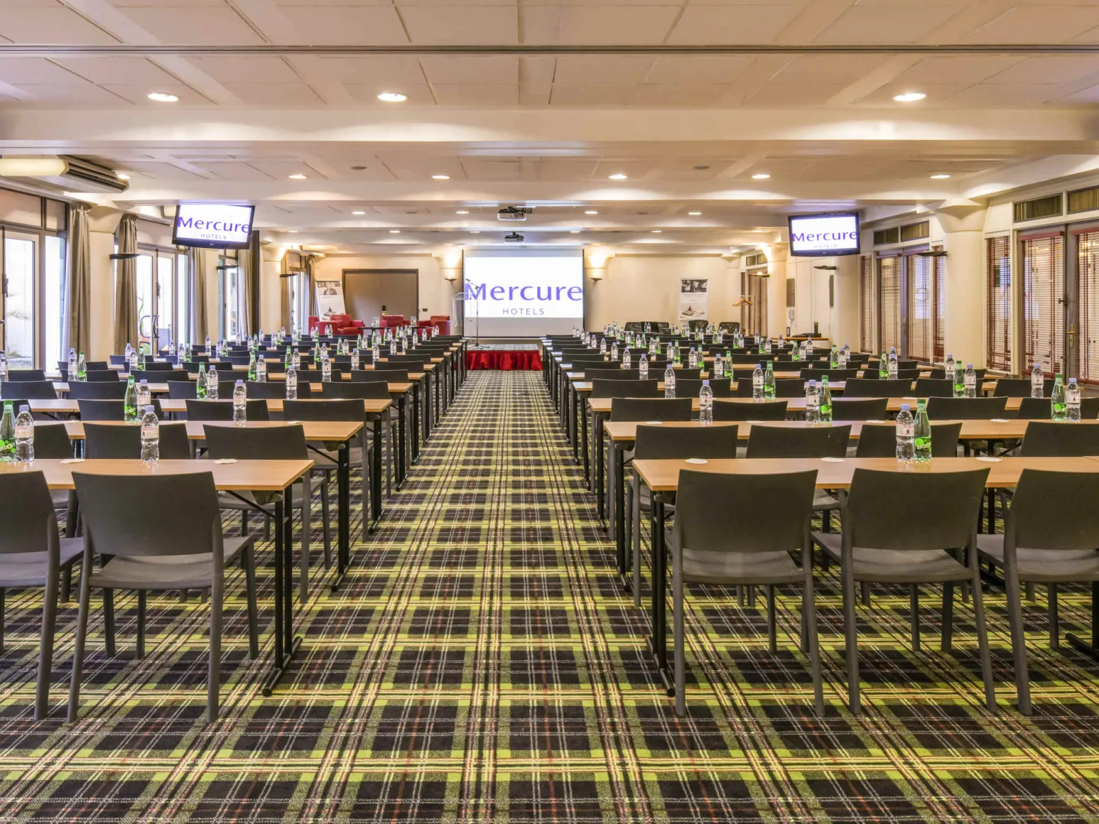 Modern conference room at Mercure Bordeaux Château Chartrons hotel with rows of tables and chairs arranged classroom style, featuring Mercure branding, projector screen, and yellow tartan carpet flooring