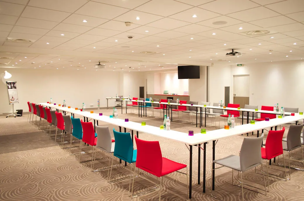 Modern conference room at Mercure Bordeaux Centre hotel with U-shaped table setup, colorful chairs in red, teal, and gray, white surfaces with refreshment cups, ceiling lights, projector, and patterned carpet flooring.