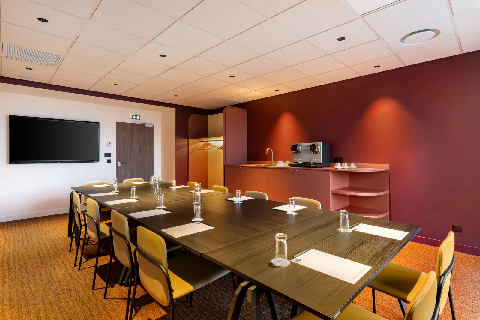 Modern conference room at Hampton by Hilton Bordeaux Mérignac with a long dark wooden table, yellow chairs, flat-screen TV on white wall, red-orange accent walls, professional lighting, and equipped kitchenette.