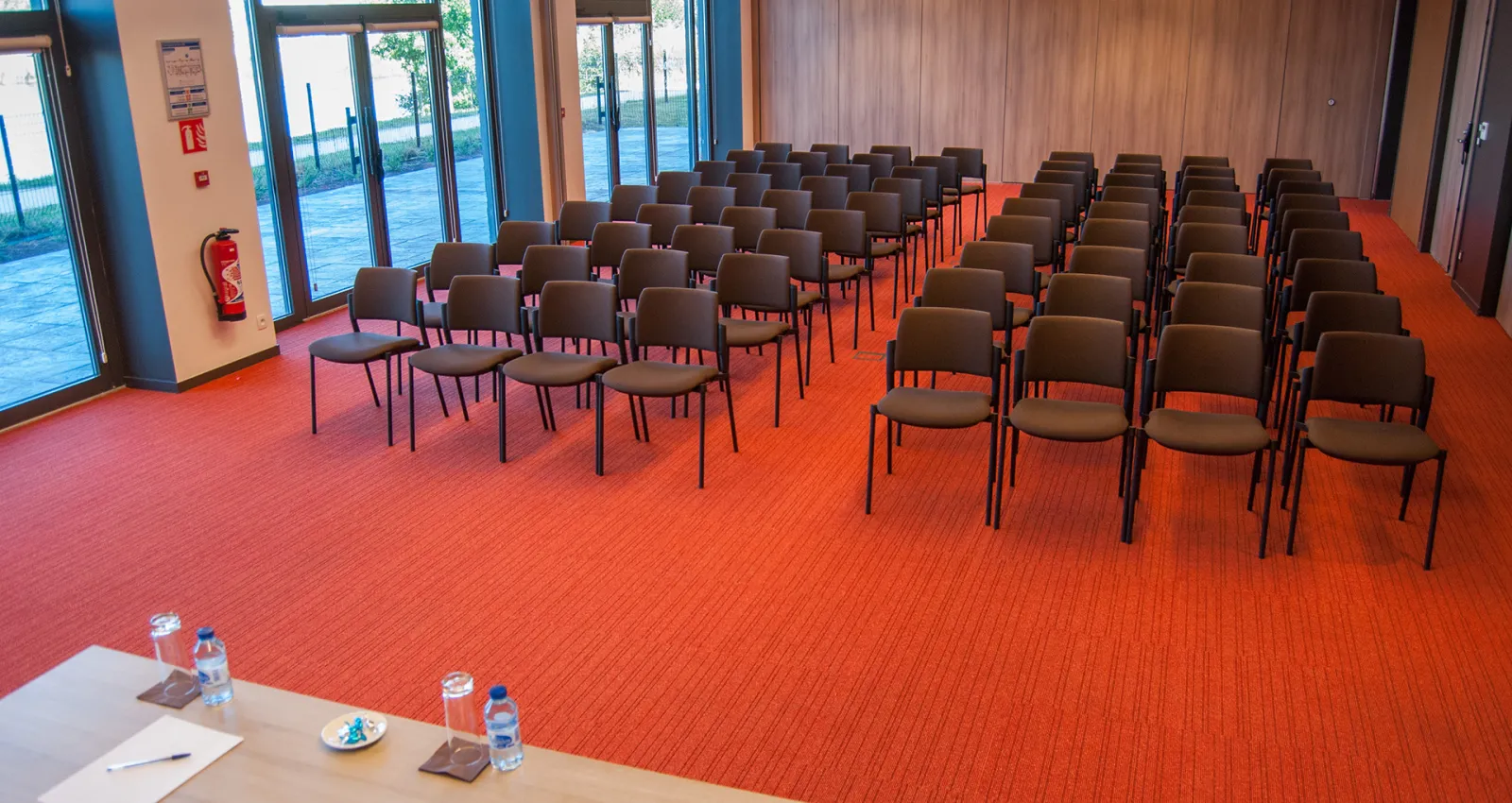 Modern conference room with brown chairs arranged in rows on an orange carpet, featuring large glass doors opening to an outdoor courtyard, wooden paneling, and a speaker's table in the foreground at Mer & Golf City Bordeaux Bruges.