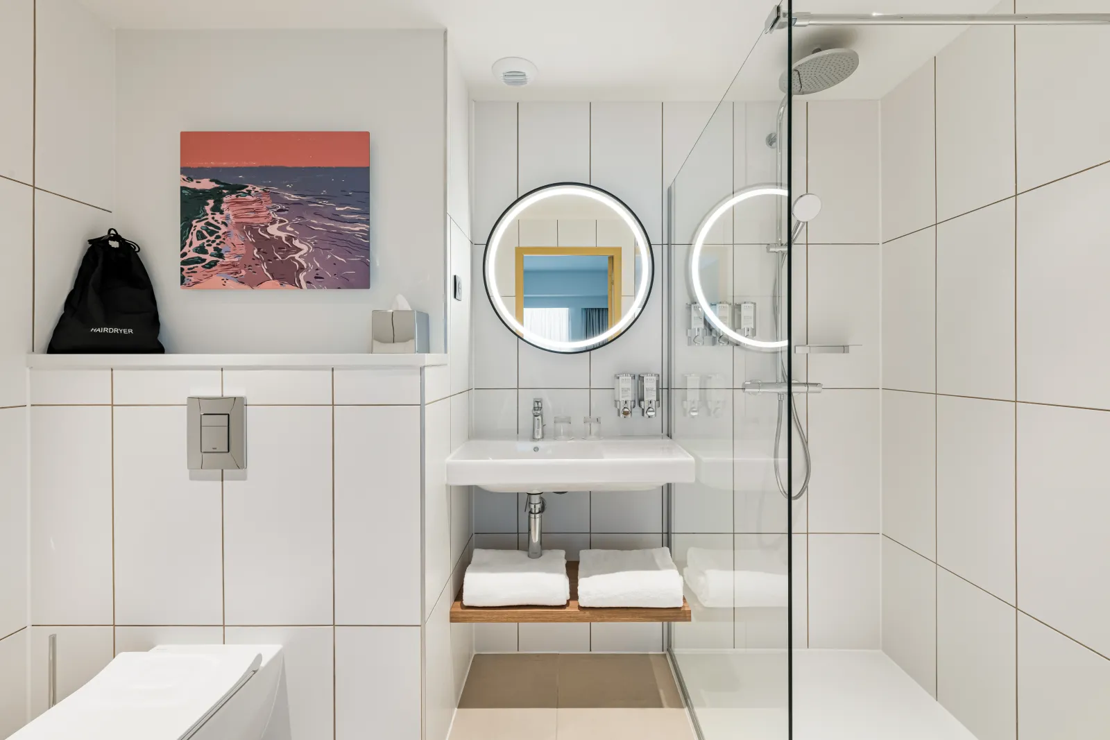 Modern bathroom with white subway tile walls, dual circular illuminated mirrors, floating wooden vanity with sink, glass shower enclosure, and colorful coastal artwork on the wall