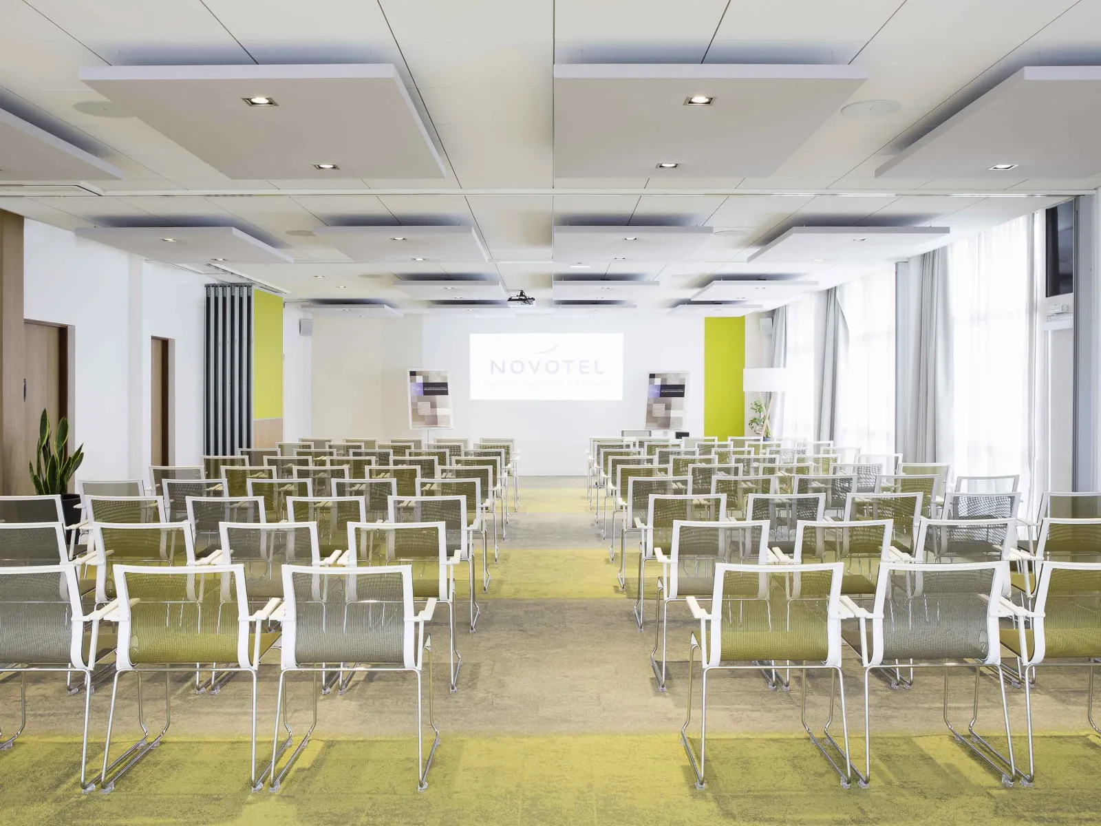 Modern conference room at Novotel Bordeaux Lac with rows of white chairs with green cushions facing a projection screen, featuring contemporary ceiling design and bright natural lighting.