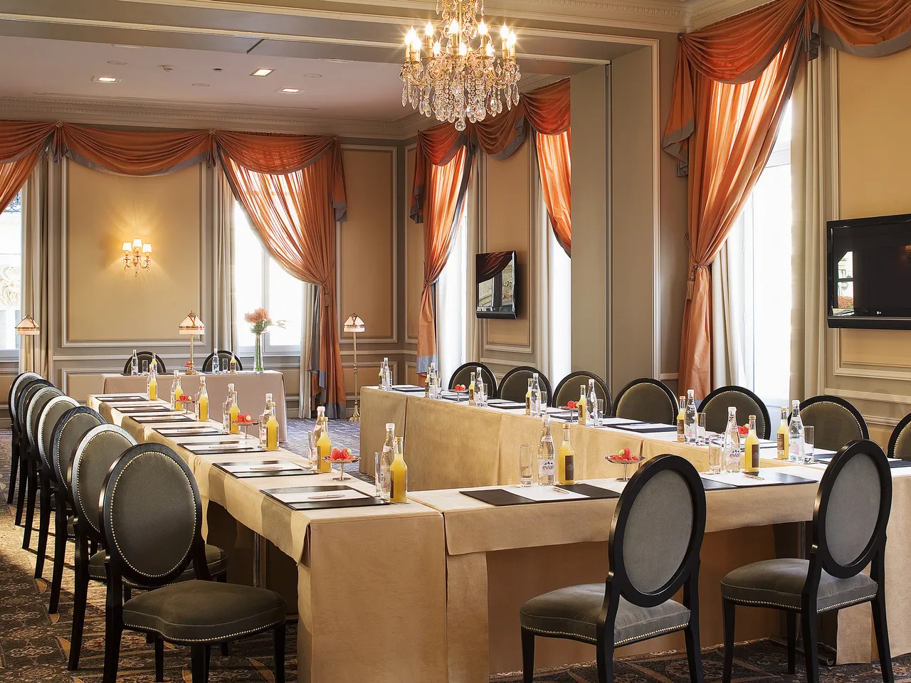 Elegant conference room with U-shaped table setup, black chairs with nailhead trim, beige tablecloths, crystal chandelier, orange velvet curtains, and tall windows with natural light at the Intercontinental Grand Hôtel de Bordeaux et Spa.
