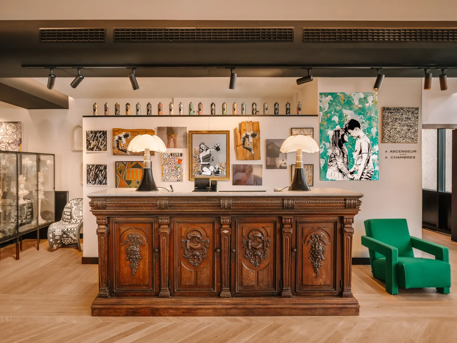 Modern hotel lobby featuring an ornate wooden reception desk with carved grape motifs, white pendant lamps, curated artwork and sculptures on the wall display, a bright green armchair, and contemporary track lighting against cream-colored walls.