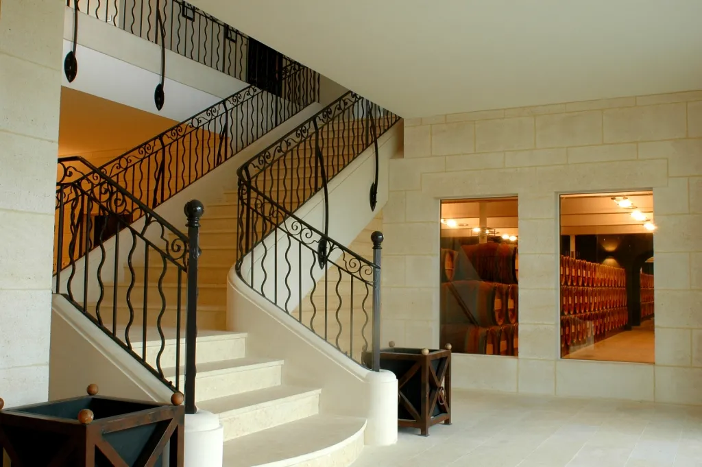 Interior staircase with wrought iron railings in a cellar at Château Léoville Poyferré in Bordeaux, featuring cream-colored steps, wooden doors with small windows, and wine barrels visible through doorways.