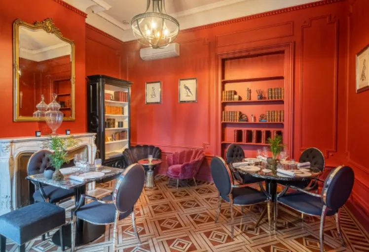 Elegant dining room at Palais Gallien Hotel & Spa in Bordeaux featuring vibrant red walls, gold-framed mirror, fireplace, blue dining chairs, burgundy velvet seating, built-in bookshelves, geometric patterned floor, and crystal chandelier.