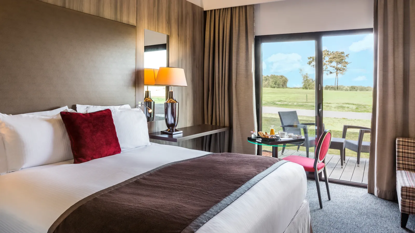 Modern luxury hotel bedroom with a queen bed, burgundy accent pillow, bedside lamp, and glass doors opening onto a terrace overlooking green countryside at Cabot Bordeaux MGallery Collection.