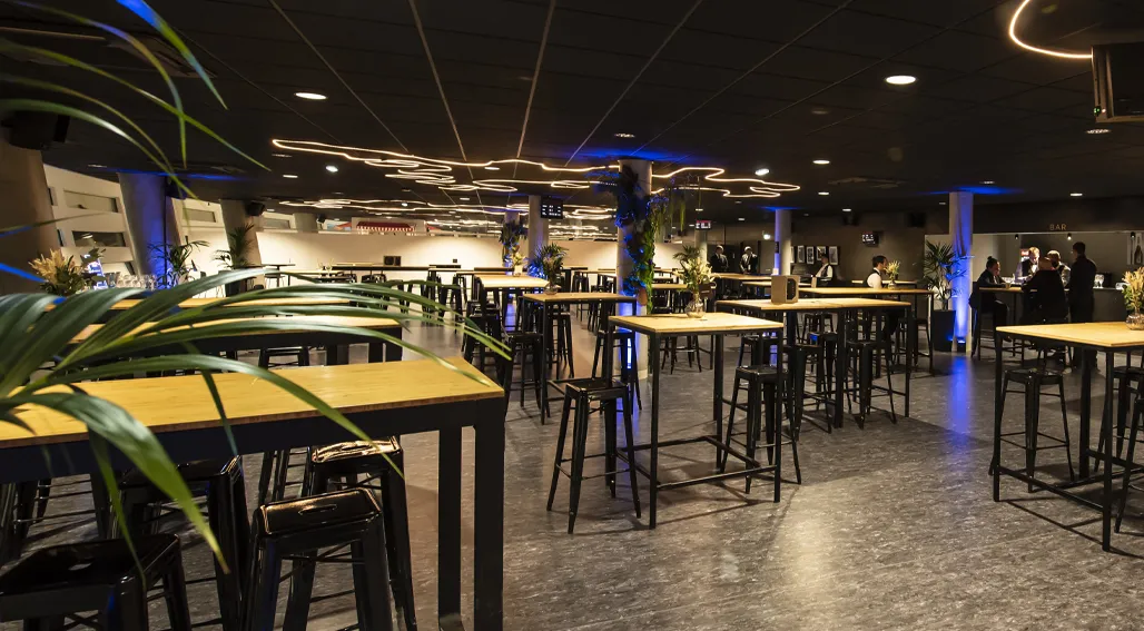Modern event space at Arkea Arena in Floirac, Bordeaux, featuring high ceilings with decorative lighting, black bar-height tables with wooden tops, black stools, green plants, and people gathered in the background during an evening event.