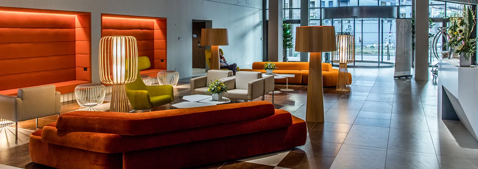Modern hotel lobby with orange and red curved furniture, wooden pendant lamps, beige armchairs, and floor-to-ceiling windows overlooking the street at Radisson Blu Bordeaux.
