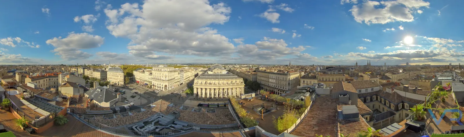 Aerial panoramic view of Bordeaux city center with neoclassical buildings, historic architecture, and urban landscape under a partly cloudy blue sky.