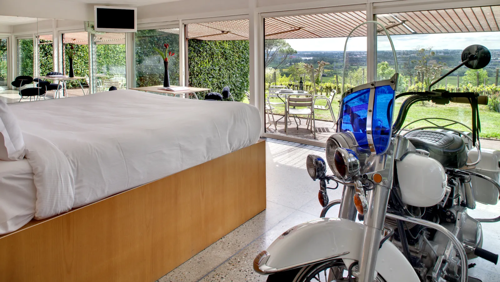Luxury bedroom at Le Saint-James hotel in Bordeaux featuring a bed with white linens and wooden frame, vintage scooters parked inside, panoramic windows overlooking vineyards, and a modern lounge area with outdoor terrace.