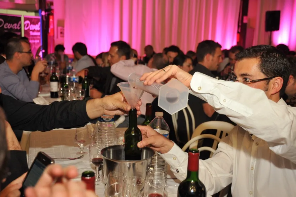 A man in a white shirt opens a wine bottle at a formal dinner event in Bordeaux, with guests seated at tables under pink ambient lighting.