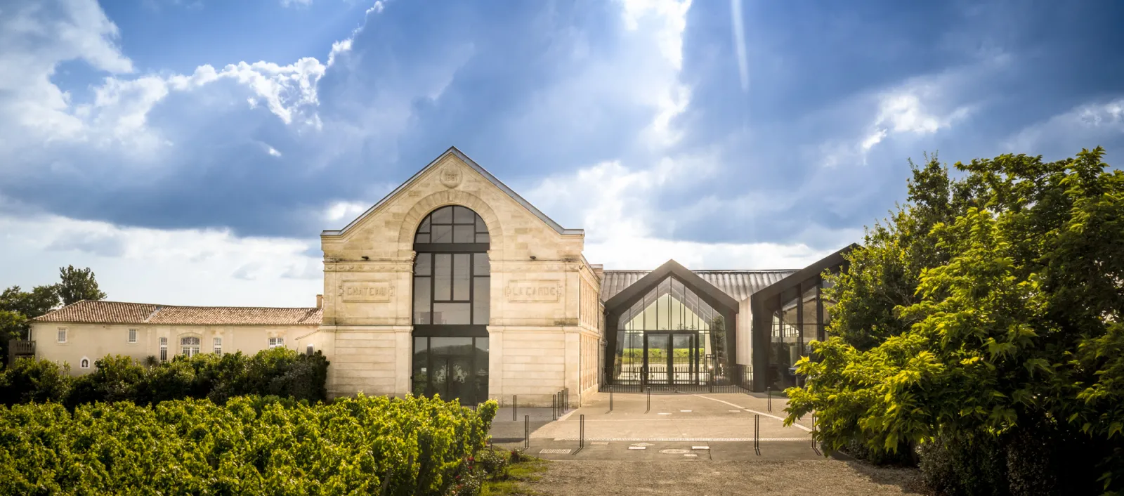 Modern wine cellar building with cream-colored stone facade and large arched window, contemporary glass extension, flanked by green hedges and vineyard buildings under blue sky with clouds