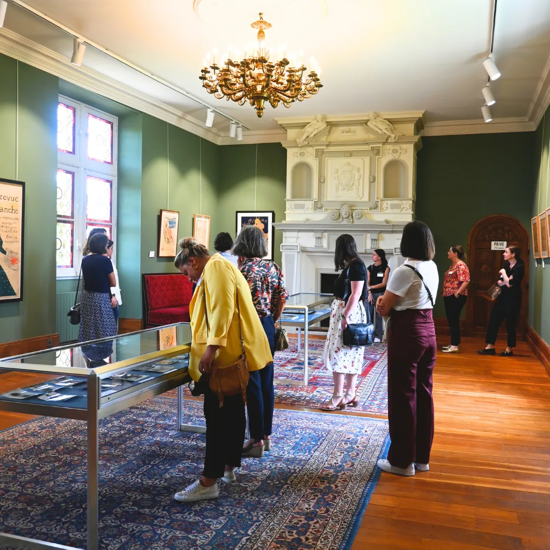 Interior of the Toulouse-Lautrec Museum in Bordeaux with visitors viewing artworks, featuring green walls, a white fireplace, ornate gold chandelier, wooden floors, and display cases.