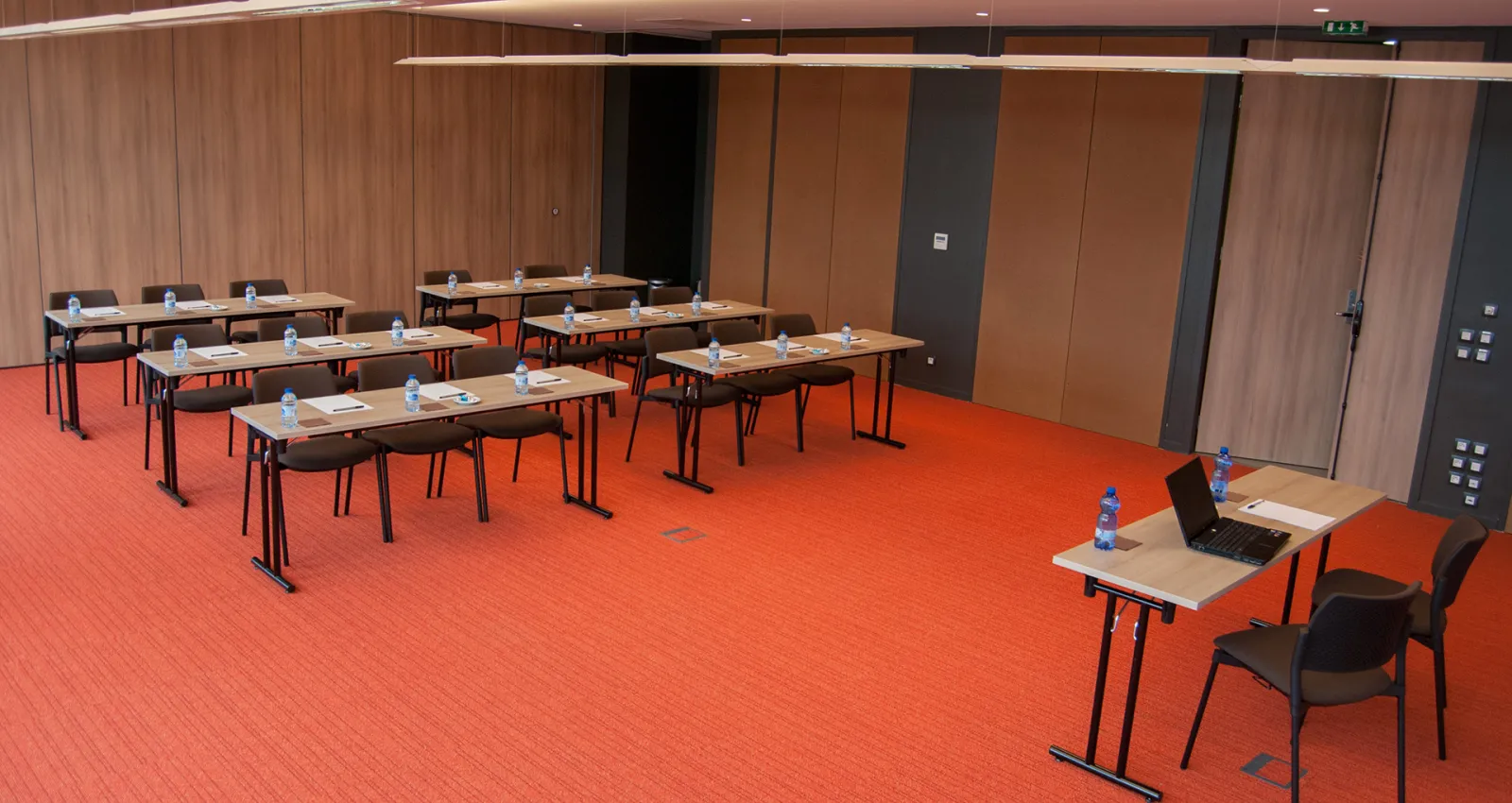 Modern conference room with multiple wooden tables arranged in rows, black chairs, water bottles, notepads, and laptops on an orange carpet with wood-paneled walls and recessed lighting.
