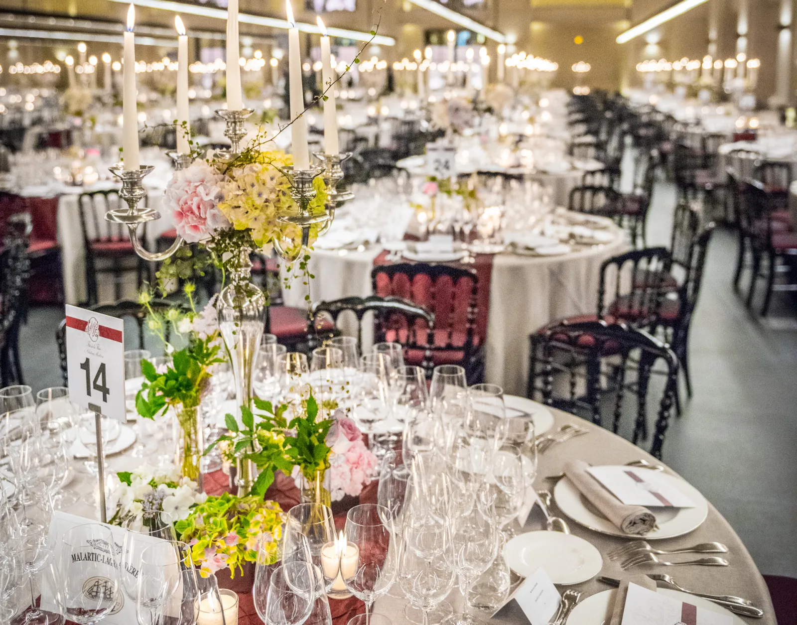 Elegant wedding reception hall with round tables dressed in white linens, decorated with yellow and pink floral centerpieces in crystal vases, burgundy chairs, place settings with white napkins, and candlelight throughout the venue.