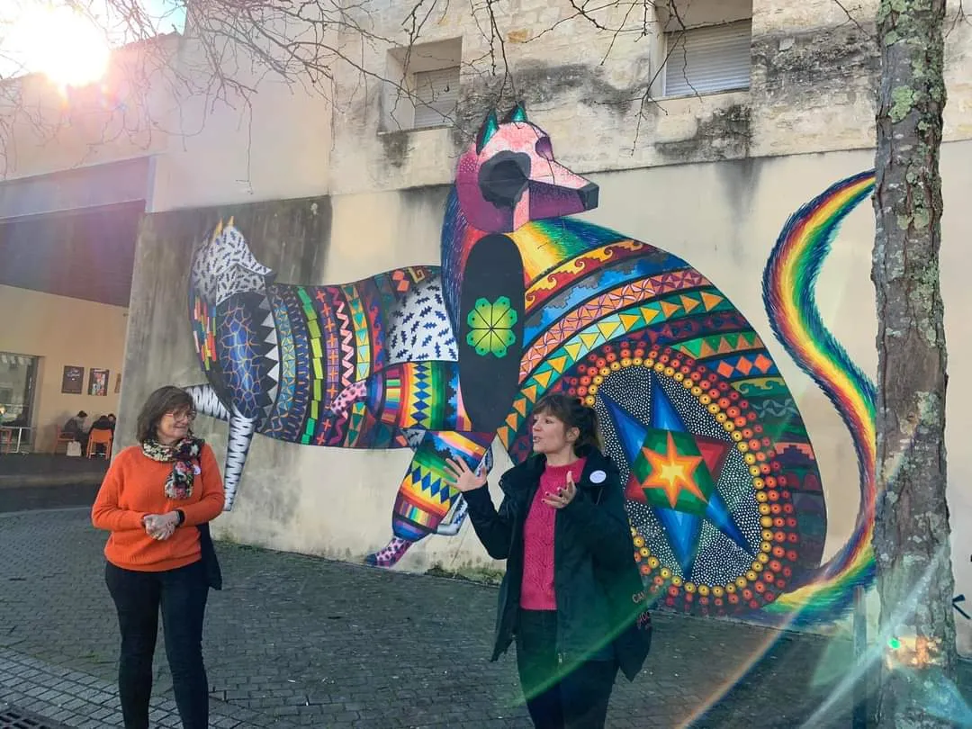 Two women standing in front of a colorful street art mural depicting a stylized animal with geometric patterns and a rainbow tail on a building in Bordeaux.