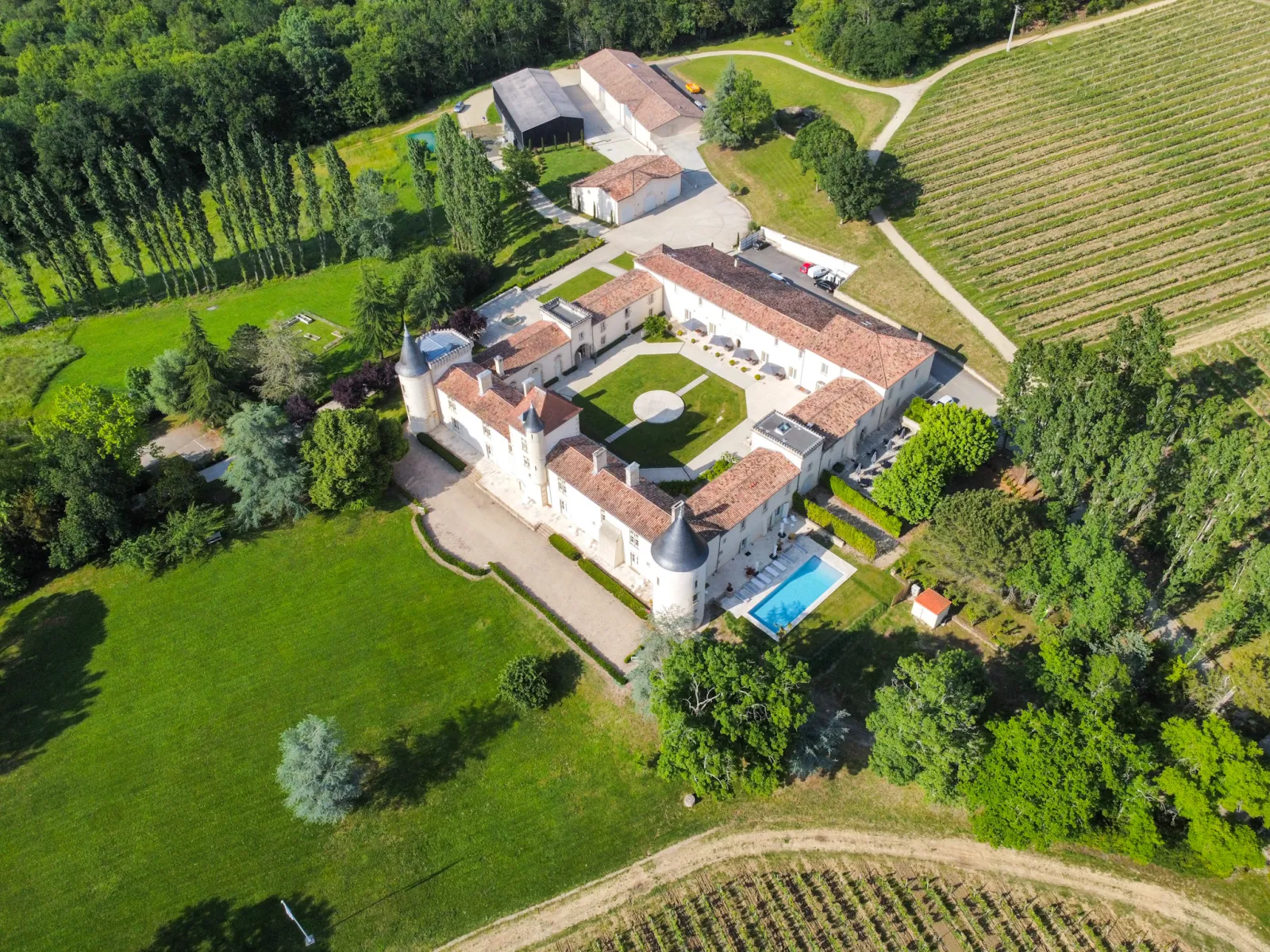 Aerial view of Château Toulouse-Lautrec near Bordeaux, showing a white stone castle with terracotta roofs, swimming pool, manicured grounds, surrounding vineyards and farmland, nestled among green vegetation and trees.
