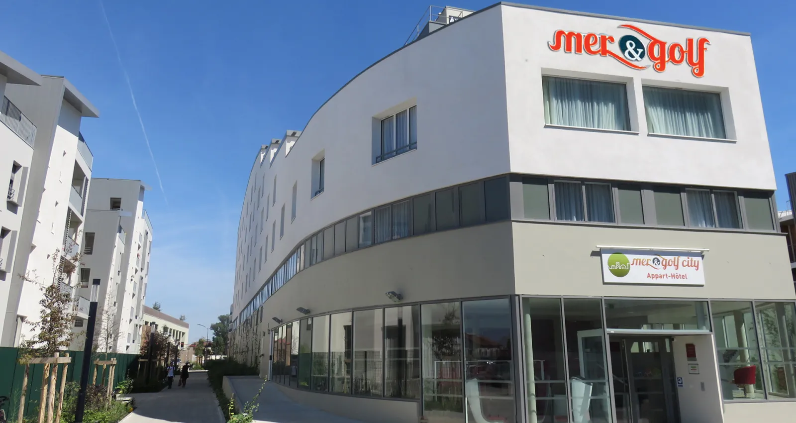 Modern white curved apartment-hotel building with 'Mer & Golf City' signage, featuring large windows and a ground floor reception area in an urban residential district.