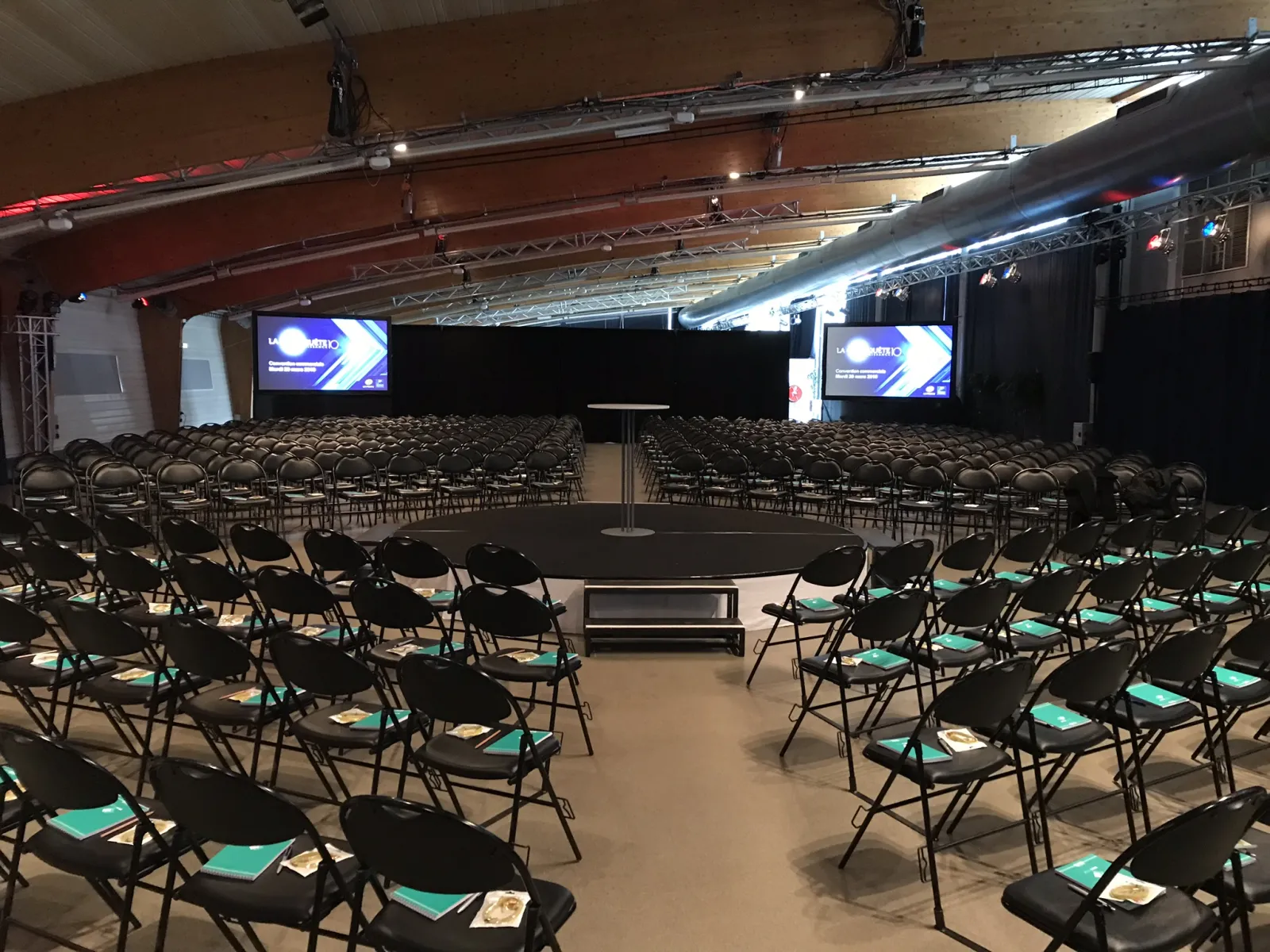 Large conference room with rows of black folding chairs arranged in a semicircular pattern, each with turquoise folders on the seat. A central podium with a microphone stands on an elevated platform. Two large screens display presentation slides on either