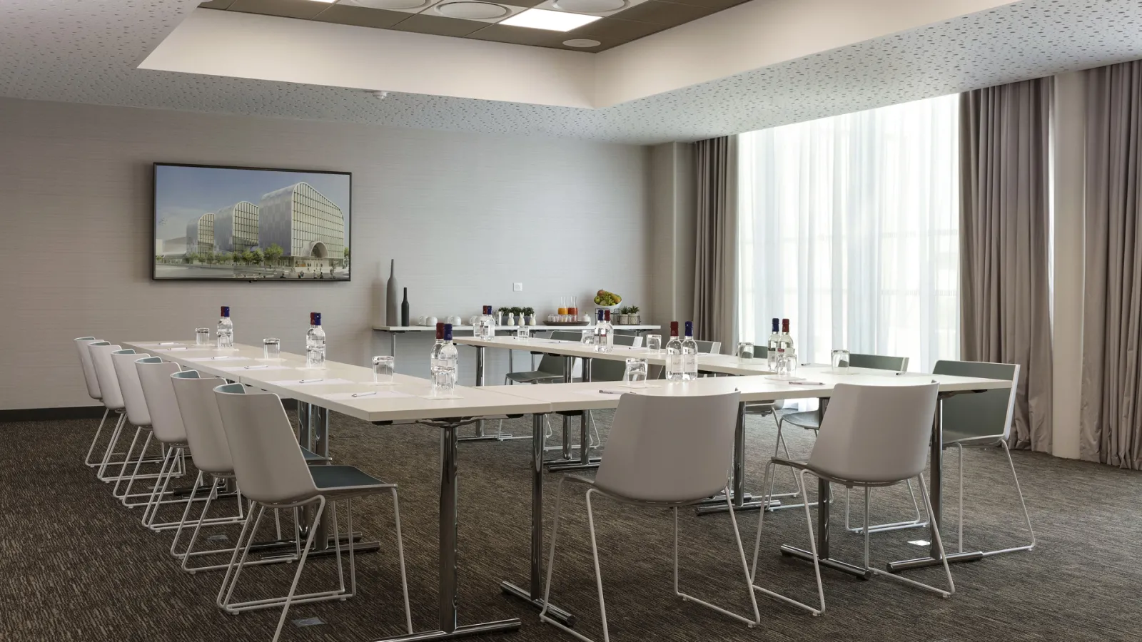 Modern conference room at Hilton Garden Inn Bordeaux Centre featuring a U-shaped table setup with white chairs, water bottles, and notepads, large windows with curtains, wall-mounted screen displaying architecture, and contemporary ceiling design.