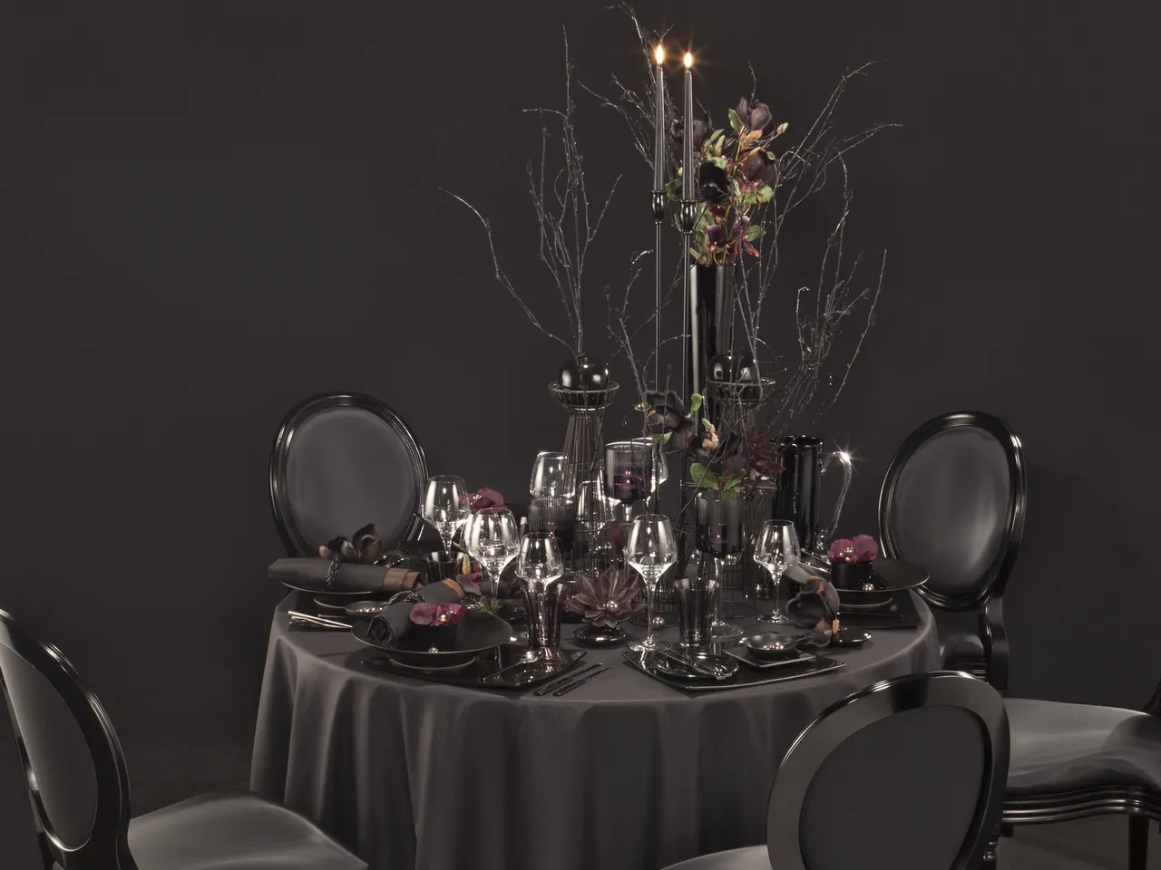 Elegant formal dining table setting with black round tablecloth, place settings on black plates, crystal glassware, black chairs with white cushions, and a tall central candelabra decorated with burgundy flowers and bare branches against a dark gray backg