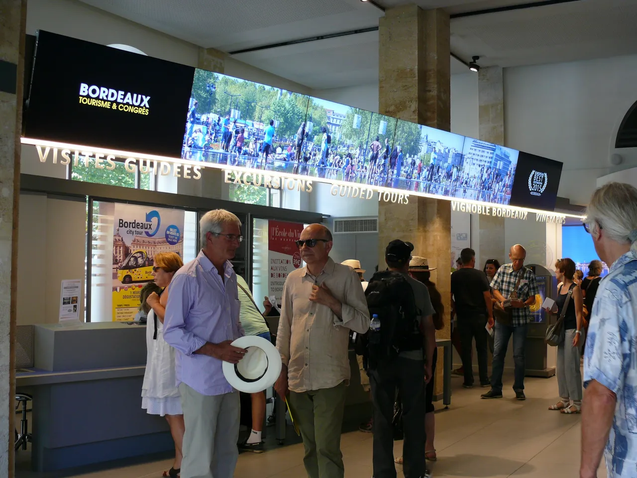 Visitors at the Bordeaux Tourism and Congress Office reception area, with a large digital screen displaying city scenes and information about guided tours and attractions