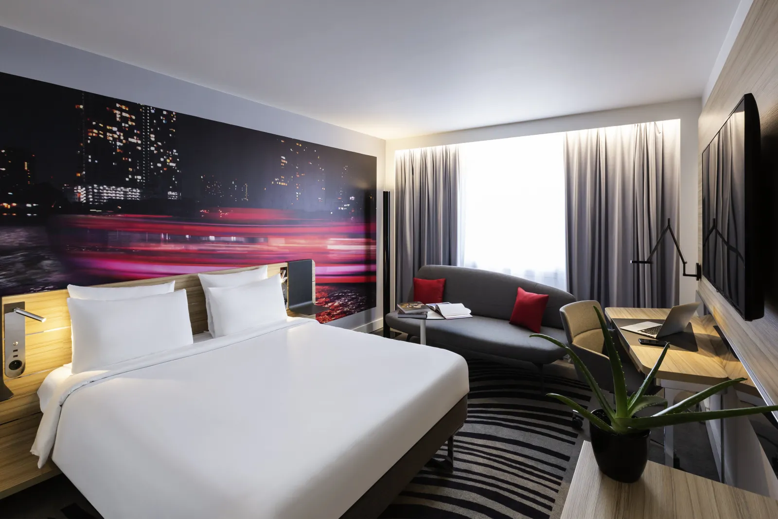 Modern hotel room at Novotel Bordeaux Centre featuring a queen bed with white linens, a gray sofa with red cushions, floor-to-ceiling windows with curtains, and a vibrant cityscape headboard with neon pink and red lights.