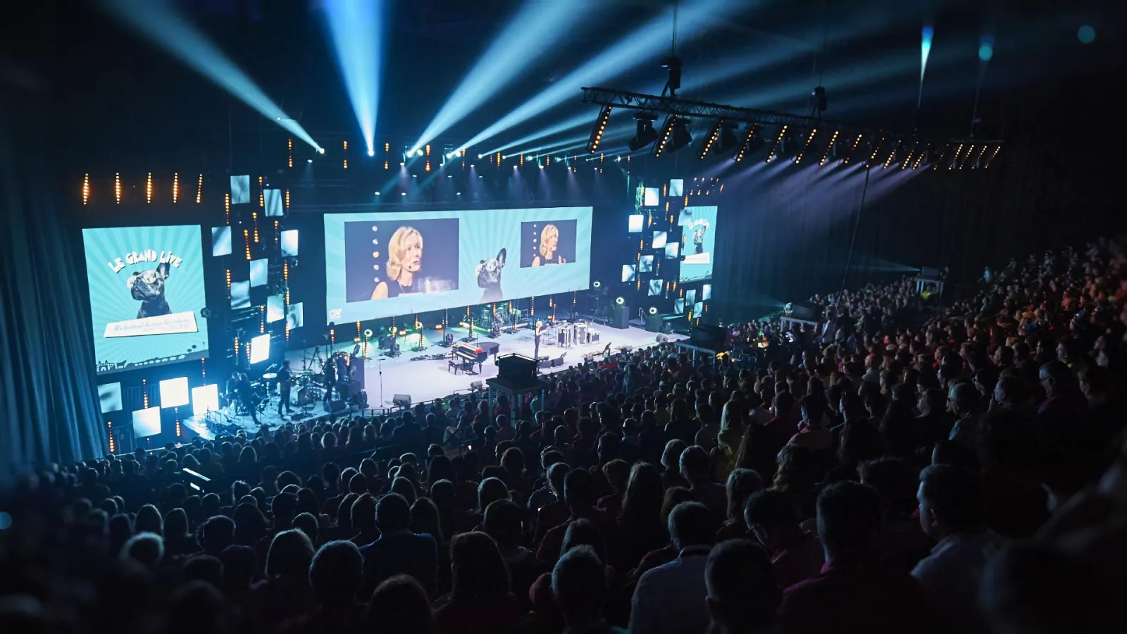 Large concert venue with thousands of attendees, elaborate blue stage lighting, multiple projection screens displaying performers, and professional sound equipment setup