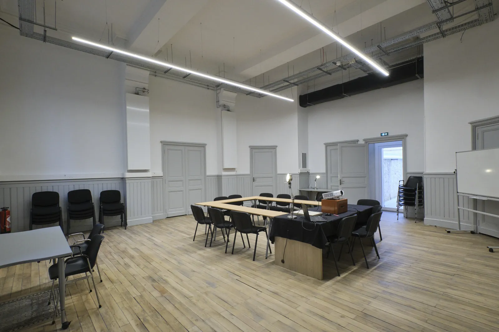 Modern conference room at ManuCo in Bordeaux featuring a large wooden table with black chairs, white walls with wainscoting, linear ceiling lights, and multiple doors leading to other spaces.
