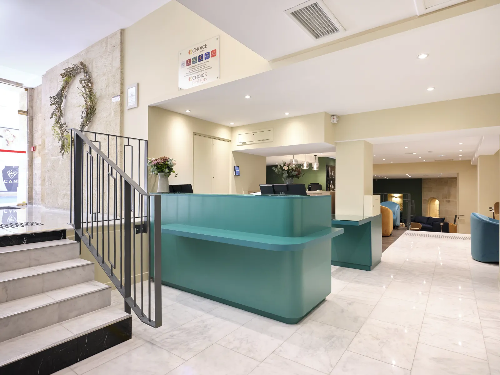 Modern hotel reception lobby with a turquoise curved desk, white ceilings with recessed lighting, marble flooring, decorative wreath on the wall, and stairs with metal railings leading to upper floors.