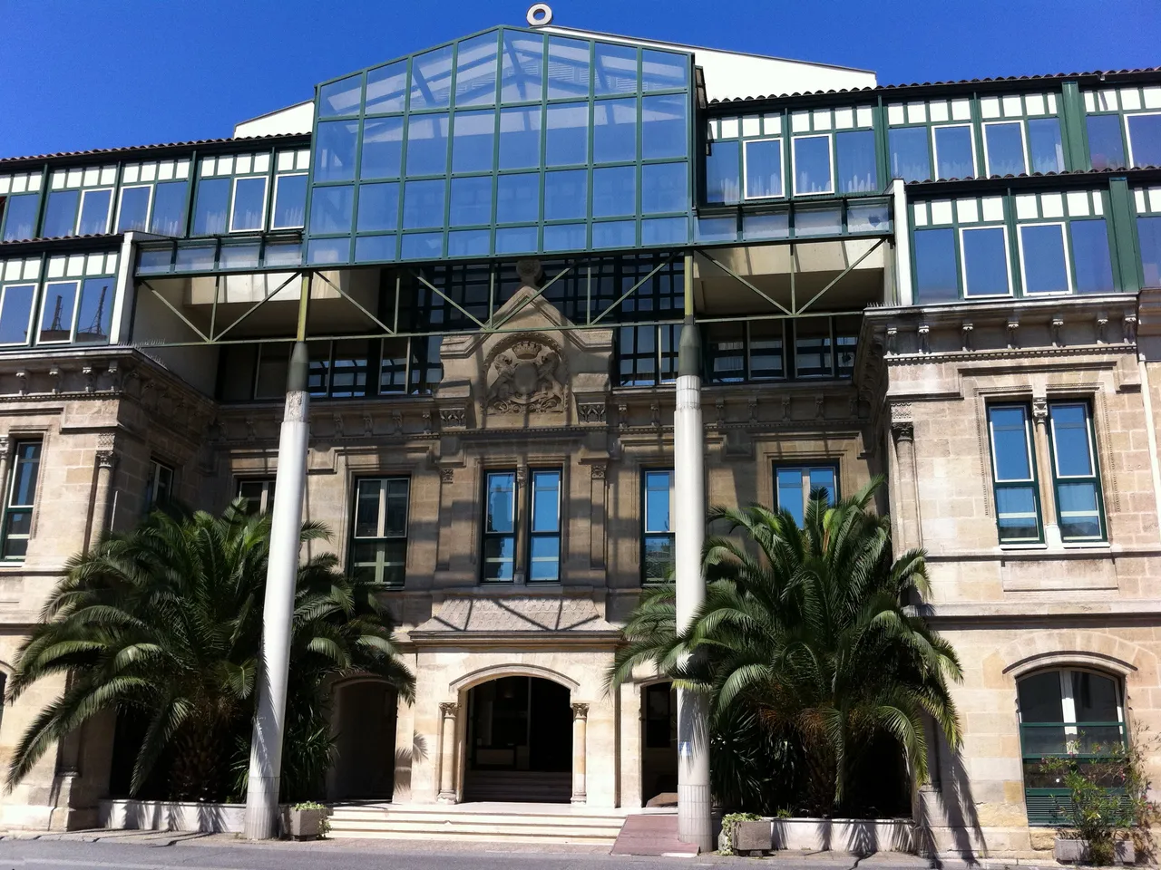 Modern glass and steel structure built atop a historic stone building facade with classical architectural details, flanked by palm trees and white columns at the entrance