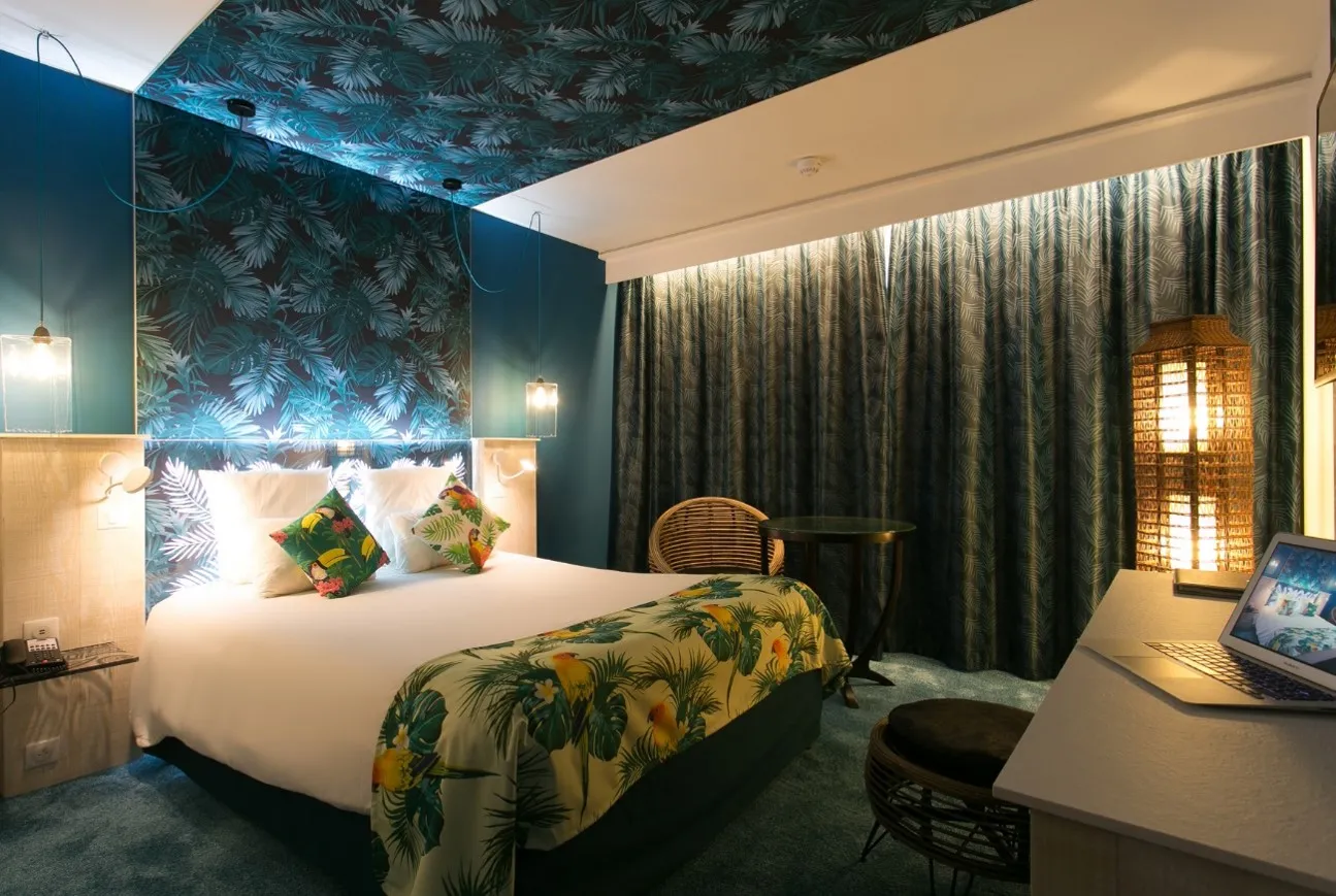 Modern hotel bedroom at Mercure Bordeaux Aéroport featuring a queen bed with tropical patterned bedding, teal botanical wallpaper accent wall, warm ambient lighting, work desk with laptop, and floor-to-ceiling curtains.