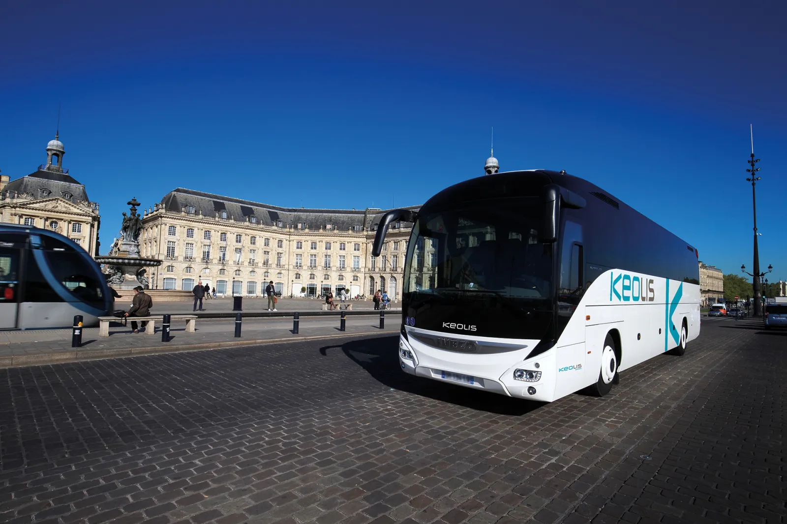 A white and dark blue Keolis coach bus parked in front of historic Bordeaux architecture on a brick-paved square, with classical buildings and a monument visible in the background under clear blue sky.