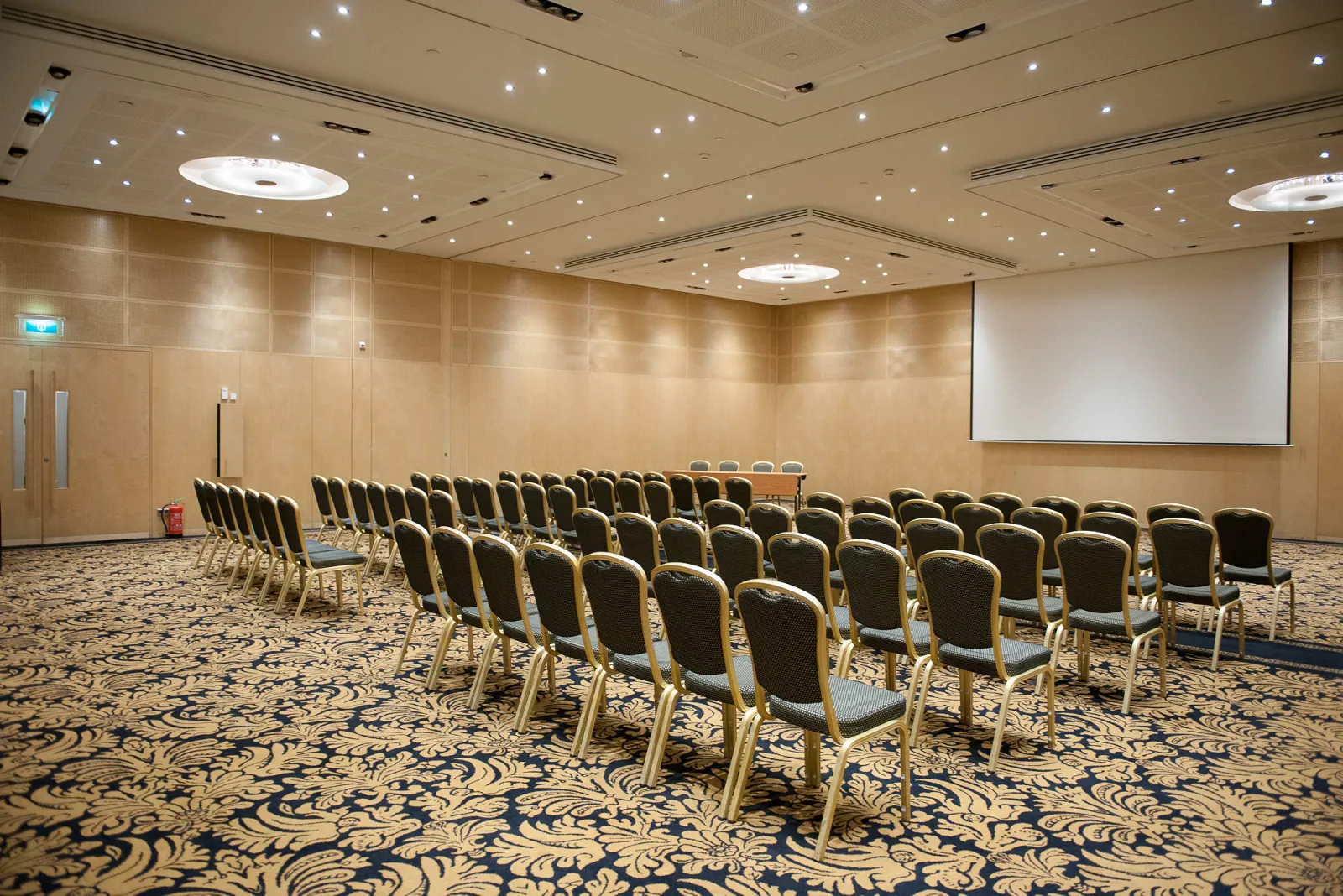 Empty conference room with rows of black upholstered chairs facing a projection screen, beige walls, modern ceiling with recessed lighting, and decorative patterned carpet at Cézame Atlantique in Bordeaux.