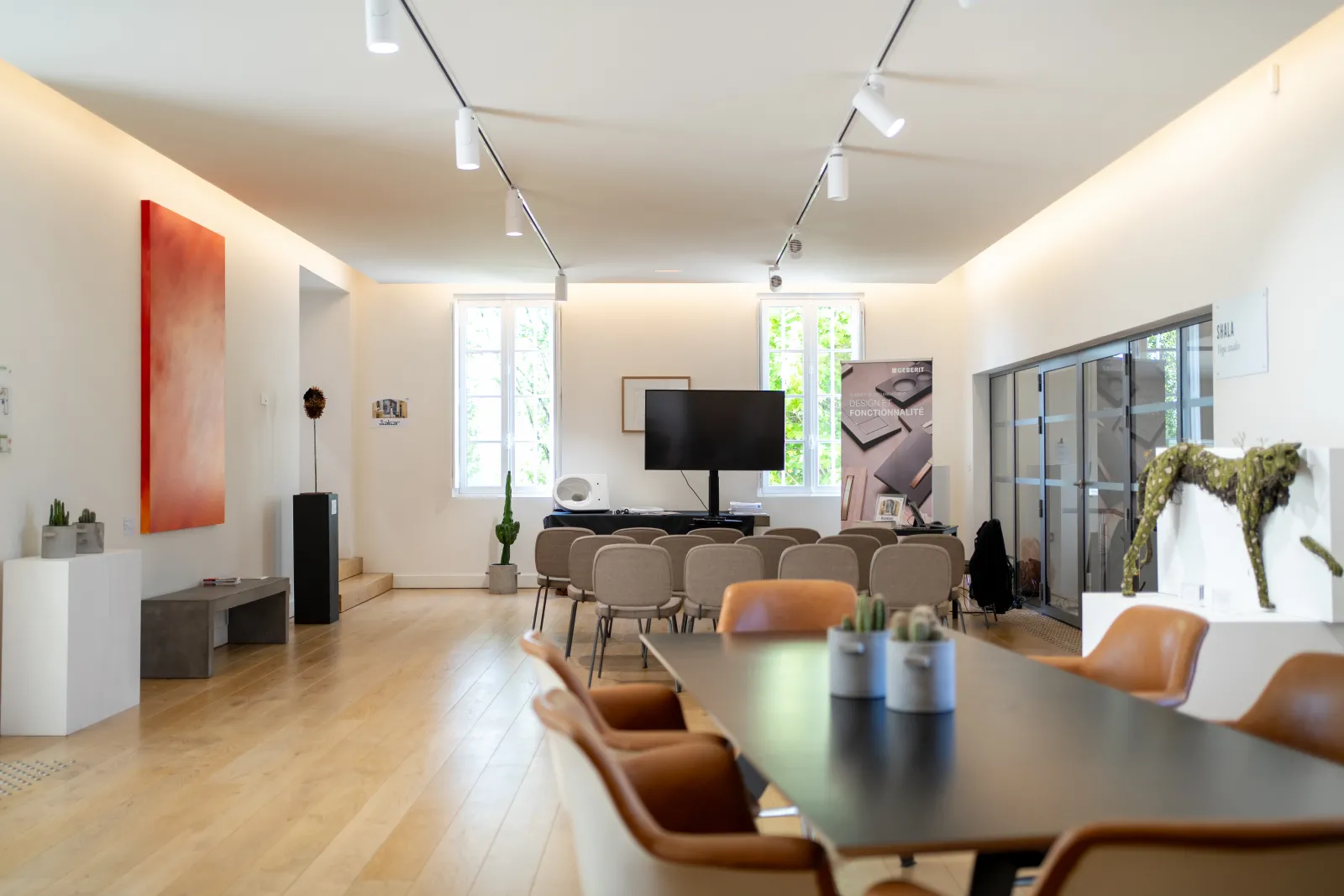 Modern conference room with wooden flooring, long table with cognac leather chairs, presentation screen, track lighting, and floor-to-ceiling windows overlooking greenery
