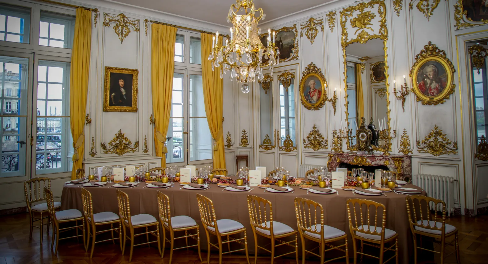 An opulent 18th-century style dining room with ornate white and gold rococo paneling, crystal chandelier, gilt-framed portraits, yellow silk curtains, and a long table set for dining with gold chairs at Maison Dulou Traiteur in Bordeaux.
