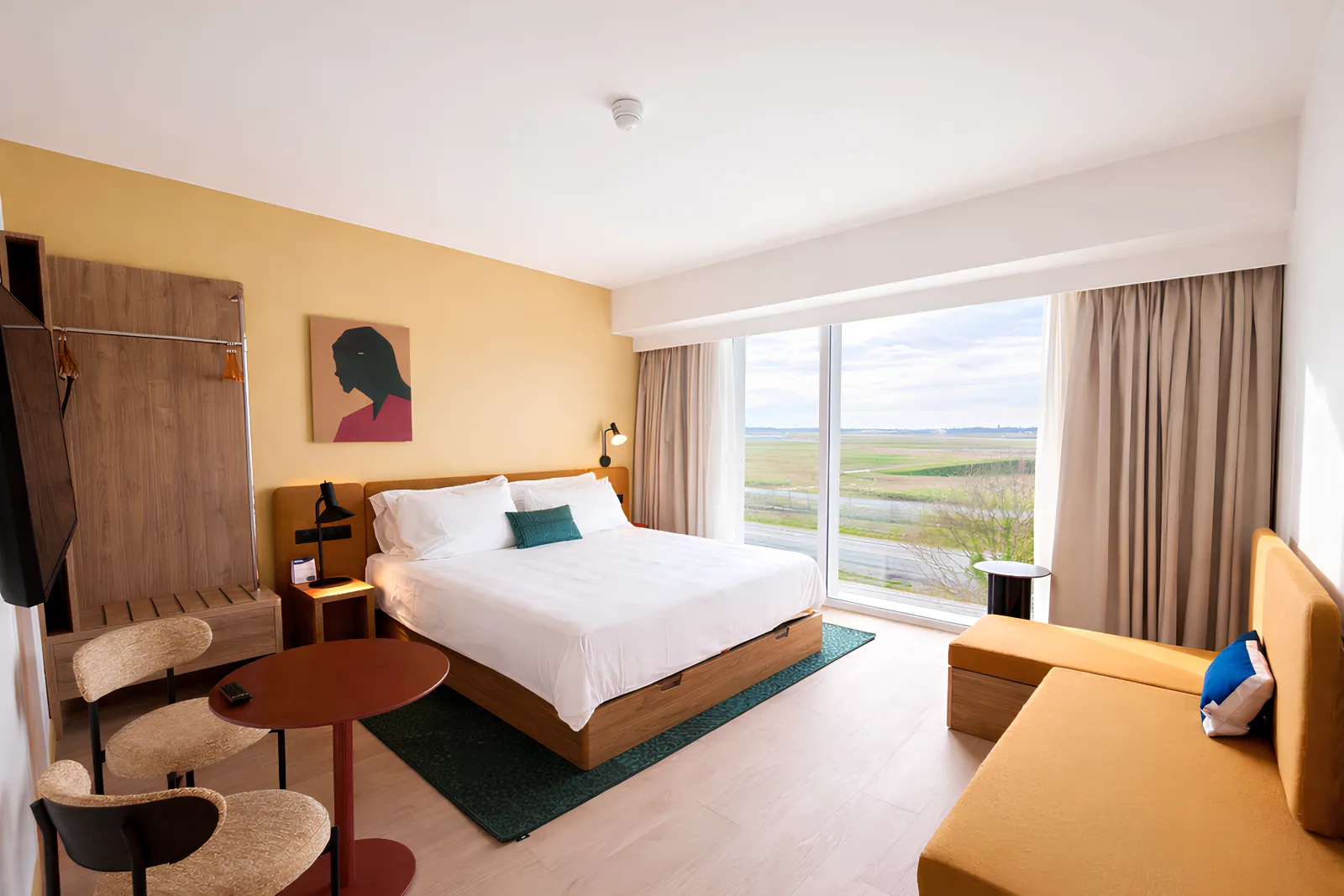 Modern hotel room at Hampton By Hilton Bordeaux Mérignac featuring a king-size bed with white linens, wooden furniture, yellow accent wall, and large windows overlooking countryside landscape with river and green fields.