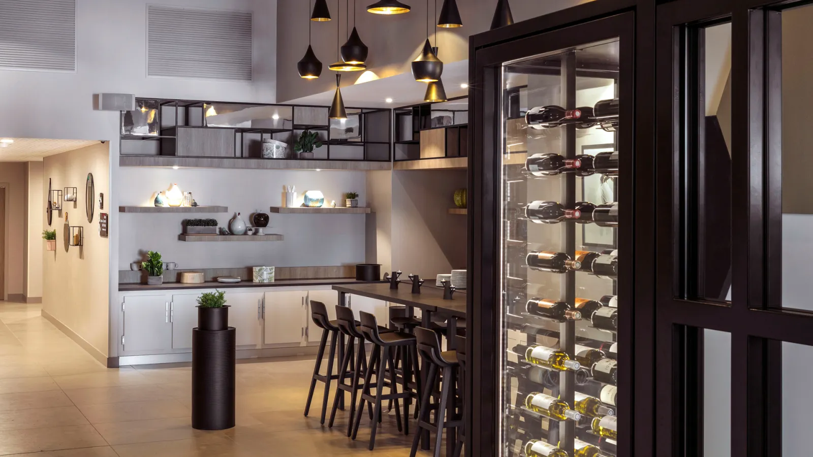 Modern hotel bar and wine lounge at Hilton Garden Inn Bordeaux Centre, featuring a sleek black and white design with pendant lighting, bar seating, open shelving with decorative items, and a glass-fronted wine cooler displaying bottles.