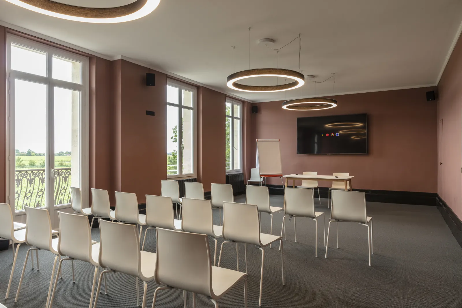 Modern conference room with cream-colored chairs arranged in rows, burgundy walls, large windows overlooking green countryside, circular pendant lights, flat-screen TV, and whiteboard.