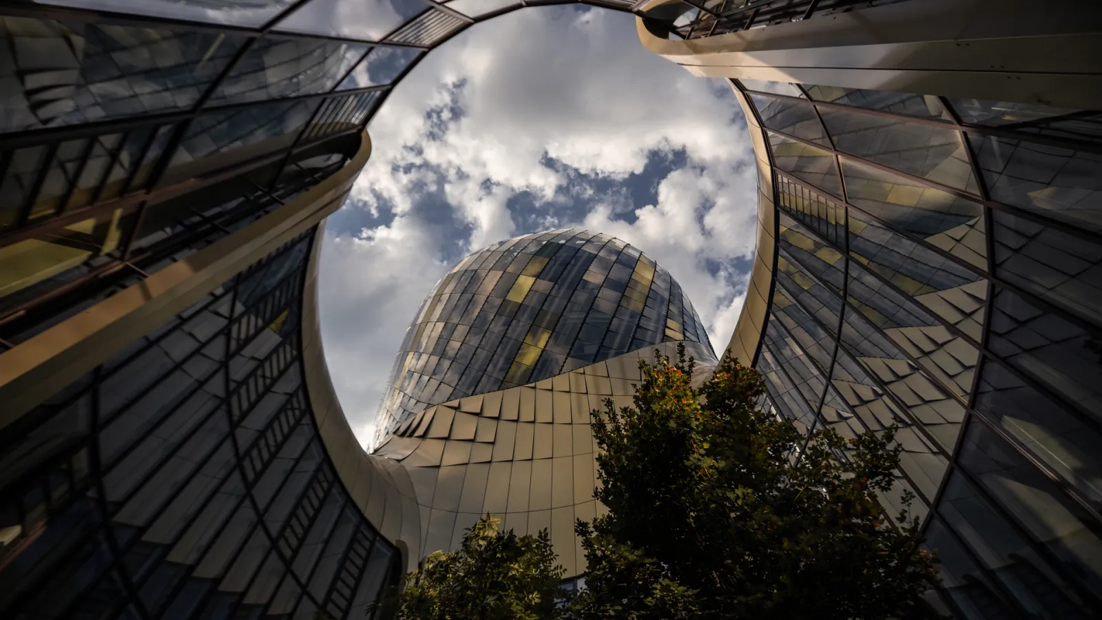 Upward view of the Cité du Vin building in Bordeaux, a modern curved glass and metal structure surrounded by contemporary architecture, with blue sky and clouds visible above.