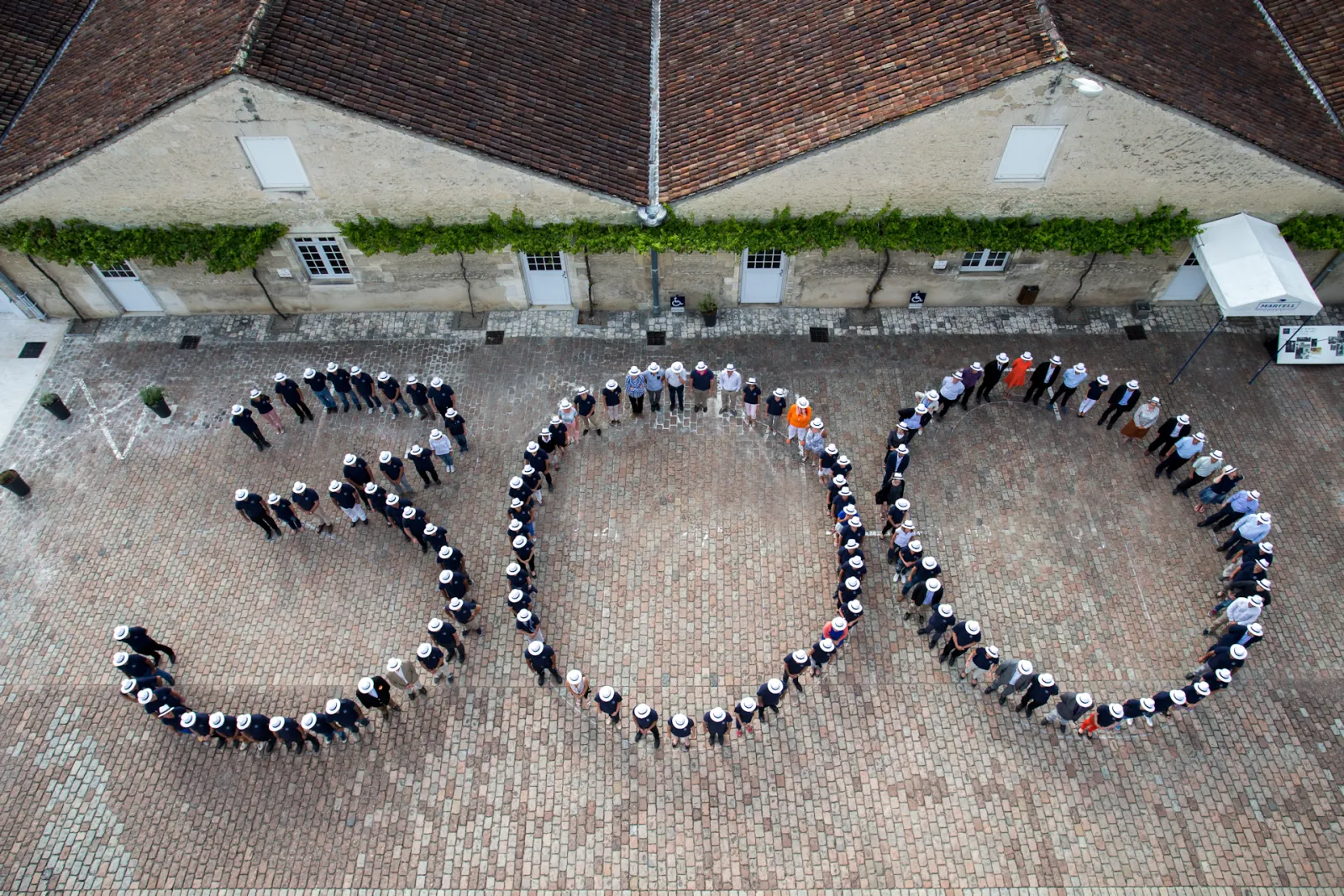 Aerial view of people arranged in a circular formation spelling '2020' in a brick courtyard in front of a historic building with ivy-covered walls in Bordeaux.