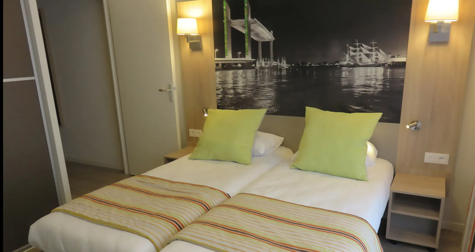 Modern hotel bedroom with double bed, green pillows, striped bedding, wall-mounted lamps, and a large window displaying a nighttime harbor view with illuminated sailing ships